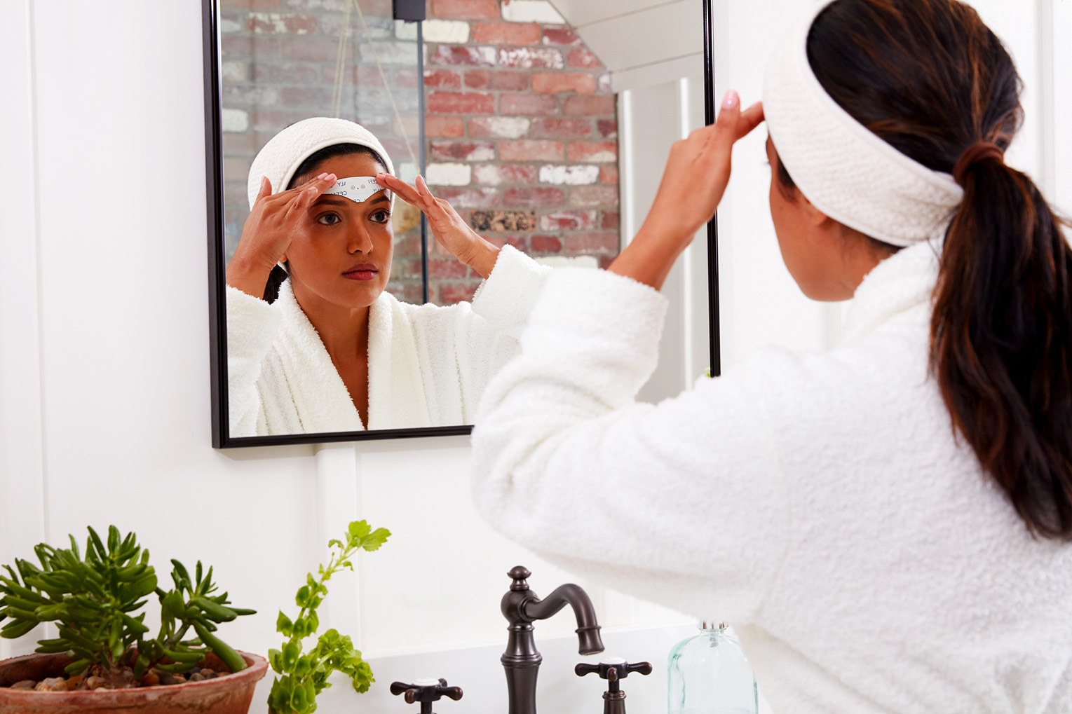 a woman is applying a CEFALY device on her forehead, as she is looking at her reflection in a bathroom mirror