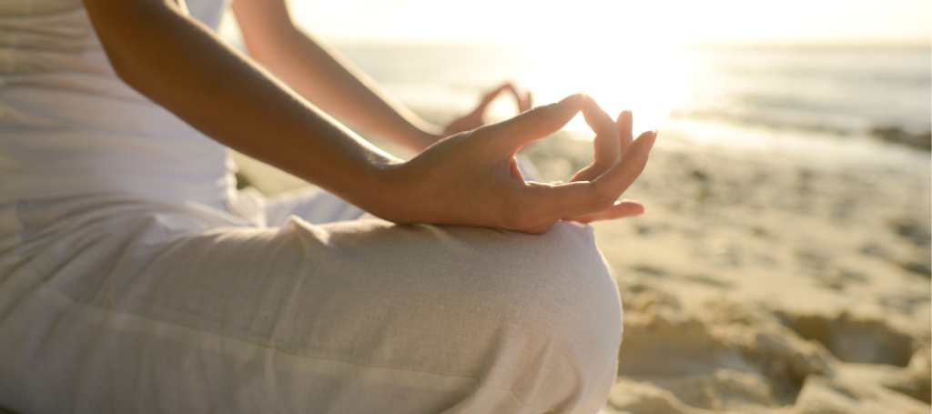 The Beginner’s Guide to Meditation for Migraine a woman meditates on the beach with her hands in a lotus position